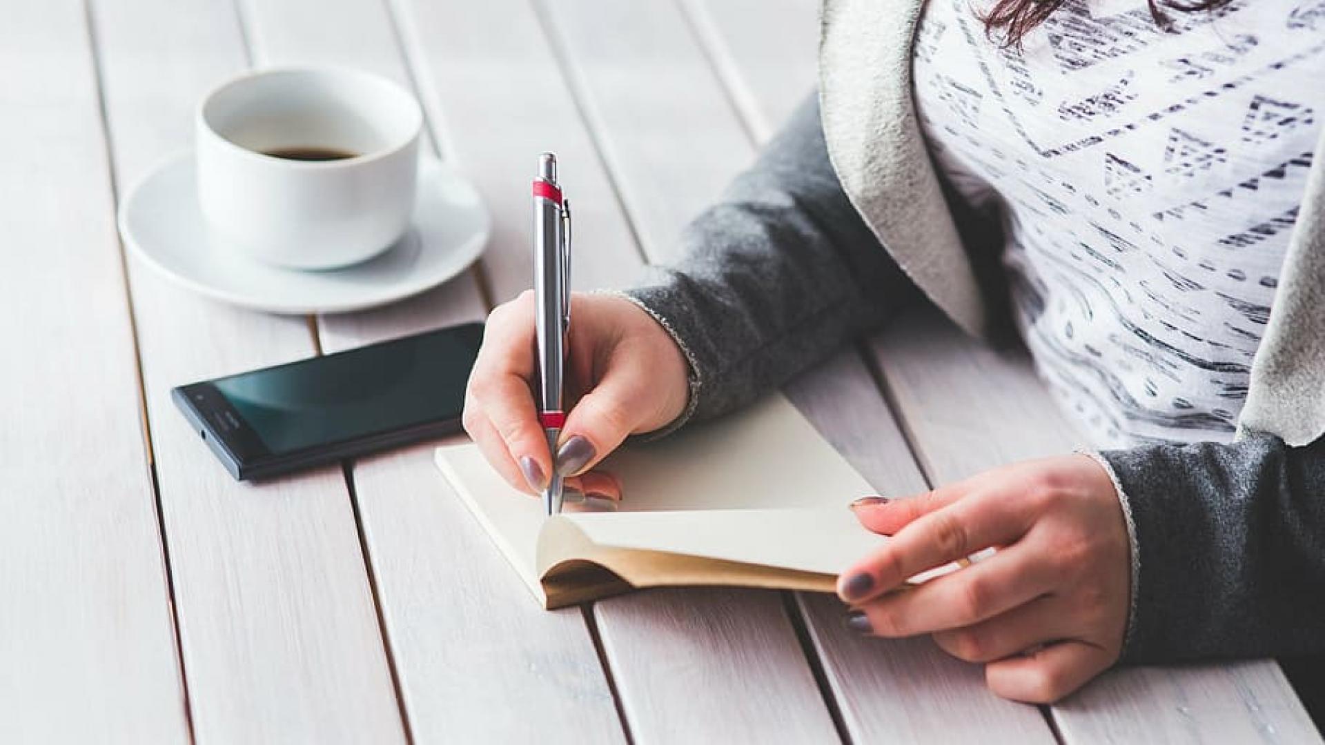 Een vrouw maakt aantekeningen aan een tafel. Naast haar ligt een smartphone en staat een kop koffie.